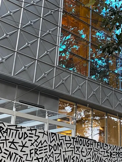 The reflection of trees on the dichroic glass of the MAP facade, with the graphic art wall below. This layering of art, architecture, and nature creates a complex and visually rich street presence.