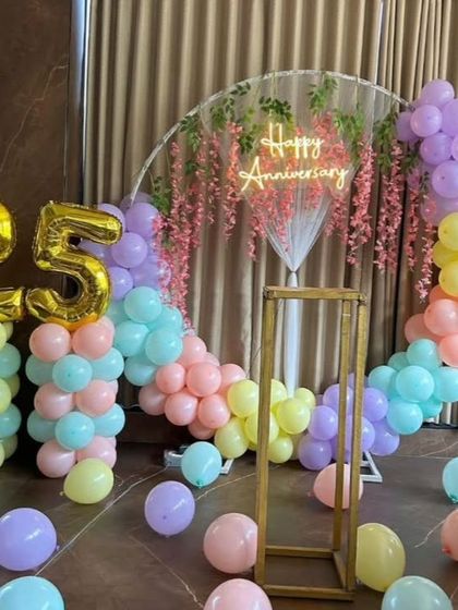 A beautiful pastel rainbow theme for a 25th anniversary. This elegant setup features a clear acrylic backdrop with a neon sign, a delicate balloon arch, and large golden foil numbers for the milestone.