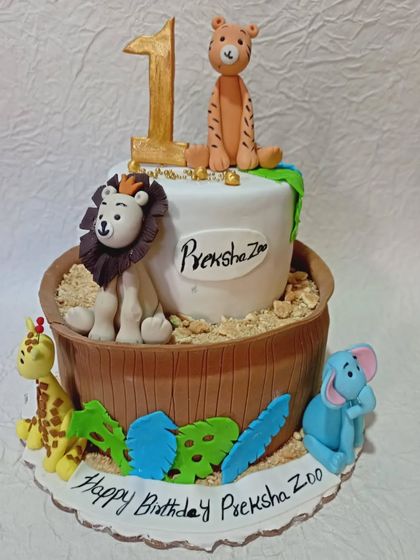 A "Preksha's Zoo" themed cake for a first birthday. This design features a variety of zoo animals like a lion, tiger, and elephant sitting in and around a wooden barrel.