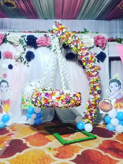 A Krishna-themed naming ceremony decoration with a hanging floral cradle and a paper flower backdrop, perfect for a vibrant celebration.