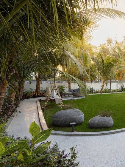 A winding pathway curves around the lawn at 'Tropical Manor', leading to a quiet seating area. The large, stone-like planters and surrounding palms create a structured yet organic feel, inviting exploration of the garden.