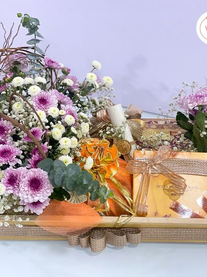 A full view of a custom wedding invitation hamper built in a rustic wooden tray. This design combines a generous floral arrangement of purple chrysanthemums with gourmet chocolate bars and a traditional scroll invite.