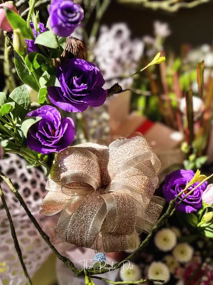 A detail shot of a shimmering, oversized bow against a backdrop of purple roses. This highlights the luxurious textures and finishes I use to ensure every executive gift feels special and celebratory.