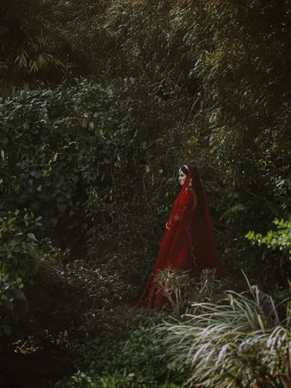 An artistic shot of a bride in a red lehenga, almost hidden within a lush, dark green forest, creating a fairytale-like image.
