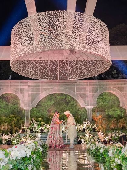 The couple during their ceremony, standing beneath the spectacular floral chandelier on the reflective aisle. The scale and beauty of the decor create an unforgettable backdrop.