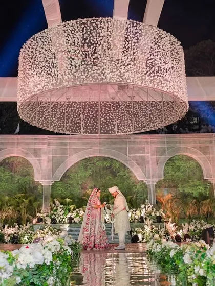 A wide shot of the couple on the stage, framed by the magnificent baby's breath chandelier and the botanical backdrop. The scale of the decor creates a truly cinematic and memorable wedding scene.