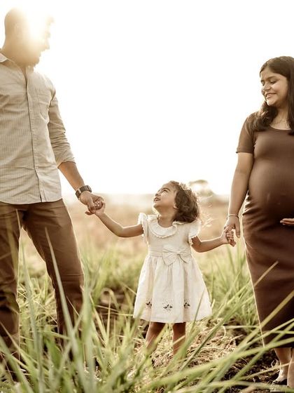 A lovely family portrait in a field of tall grass. The parents hold their daughter's hands, and she looks up at them with love, all three connected and happy.