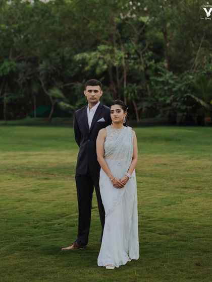A formal portrait of the couple in a lush green field. The bride's light blue saree and the groom's dark suit create a beautiful contrast against the natural backdrop.