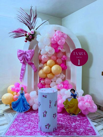 A truly magical Disney Princess theme for Taara's first birthday. This setup features a stunning balloon cascade in pinks and peaches flowing out of a white arched structure, complete with princess cutouts.