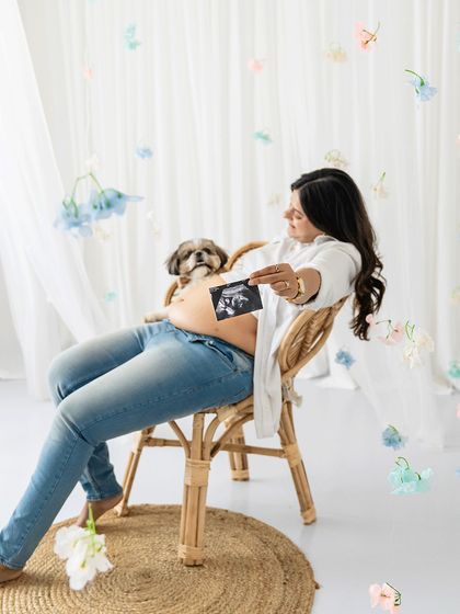 A beautiful portrait from a studio maternity session. An expectant mother holds her Shih Tzu and the sonogram, surrounded by delicate hanging flowers, creating a dreamy and heartfelt announcement.