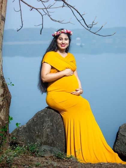 A serene lakeside portrait. The mom-to-be, wearing a yellow gown and floral crown, looks peacefully out over the water, creating a calm and beautiful image.