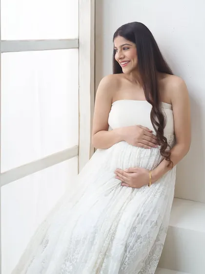 A happy, candid moment captured by the window. The mother-to-be's joyful smile and the bright, natural light make this a heartwarming and authentic portrait.