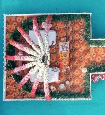 An aerial top-down view of a unique floating mandap. The structure is built over a pool, with a vibrant printed floor and a sunburst pattern of floral garlands creating a spectacular visual.