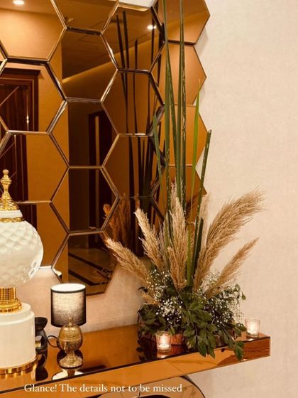 A detail shot from a Diwali decor setup. A beautiful arrangement of pampas grass and baby's breath sits on a gold console table in front of a honeycomb mirror, creating a chic and festive corner.