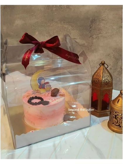 A beautiful pink Eid bento cake in its clear gift box. The cake is decorated with a crescent moon and a cute illustration, tied with a festive red ribbon.