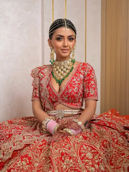A seated portrait capturing the grandeur and grace of the bride. The rich red lehenga and intricate jewelry are balanced with makeup that is both glamorous and refined, perfect for the main event.