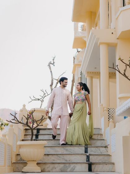 A grand, wide shot of the couple at their Welcome Lunch venue. Their pastel outfits look stunning against the architecture, creating a regal and romantic image.