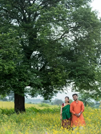A wide shot of an expecting couple in a field of yellow flowers, with a large, majestic tree behind them. Their traditional Indian outfits, a green saree and orange kurta, add a vibrant touch to this classic outdoor maternity portrait.