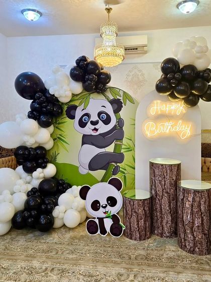 A fun panda-themed birthday party setup. The decor features a large panda backdrop, a neon "Happy Birthday" sign, and a black and white balloon arch.
