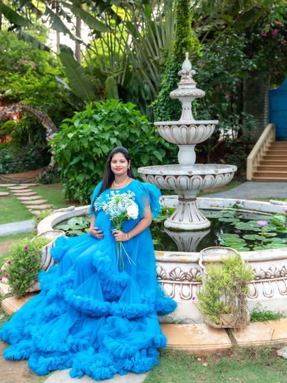 A serene portrait of a mom-to-be in a blue ruffled gown, sitting by a garden fountain.