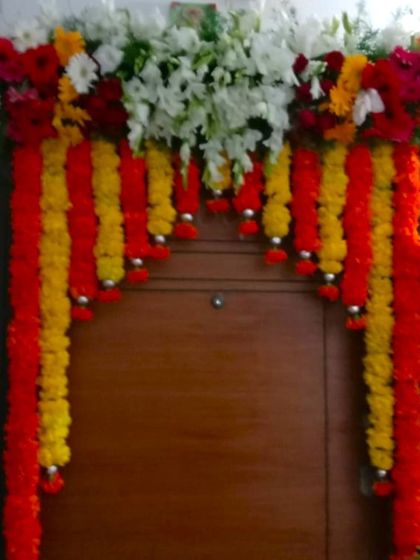 A front door decoration using red and yellow artificial flower garlands. The top is accented with white flowers and silver bell hangings.