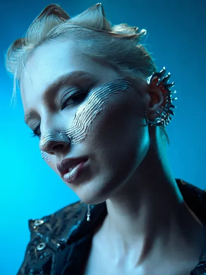 A close-up of a futuristic look featuring textured silver makeup across the nose and cheeks and spiky, sculpted hair. The cool blue lighting enhances the metallic sheen and the edgy, sci-fi vibe of the entire concept.