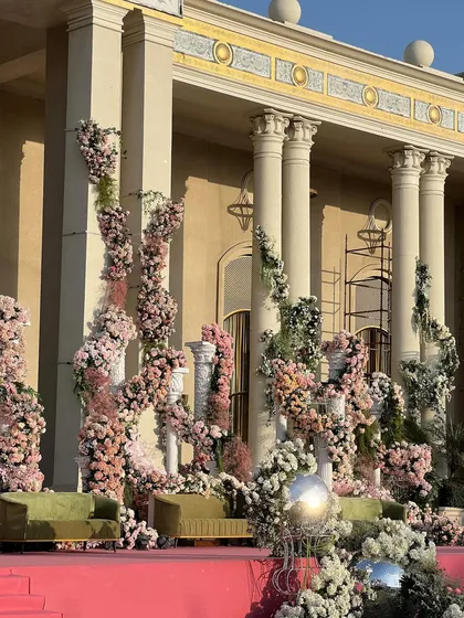 A unique stage backdrop for a cocktail event, featuring pillars adorned with cascading pink and white florals against the grand architecture of the venue. The design blends romantic floral artistry with a sense of classical grandeur.