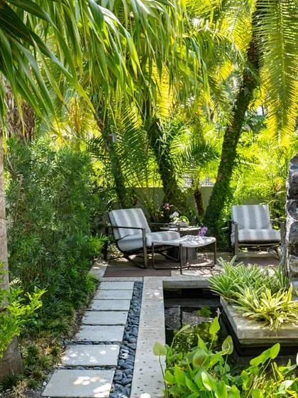 A secluded seating area next to the pond, surrounded by palm trees and lush undergrowth. We create private nooks within larger gardens for quiet relaxation.