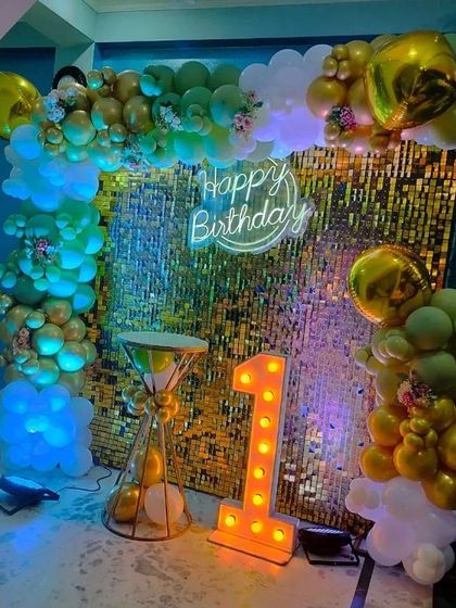A first birthday setup with a gold shimmer wall, a light-up number '1', and a green and gold balloon arch.