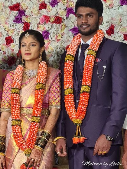 A wedding couple standing before a floral backdrop. The bride's makeup is soft and elegant, letting her natural features and beautiful saree shine.