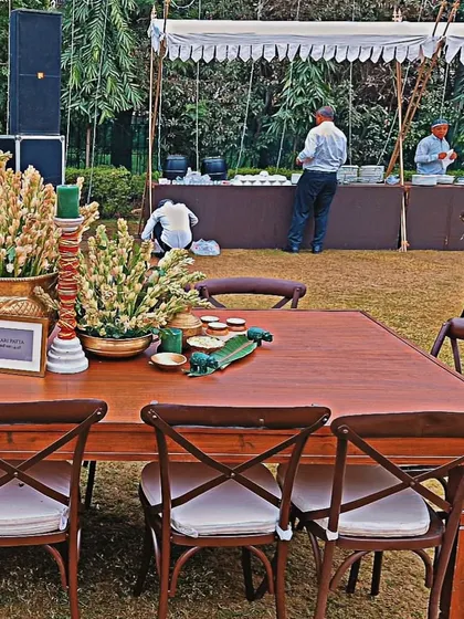 A classic outdoor dining setup with rustic wooden tables and cross-back chairs. The centerpiece of white tuberoses in a traditional brass pot adds a touch of Indian elegance to the simple, clean aesthetic.