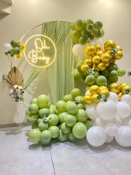 For a chic and modern baby shower, we offer gender-neutral themes like this green and gold setup. It features a circular hoop with an "Oh Baby" neon sign, draped fabric, and an organic balloon garland in olive green, white, and chrome gold.