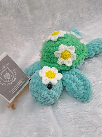 A single, sweet turtle plushie made with soft blue and green yarn and adorned with a daisy on its head and shell.