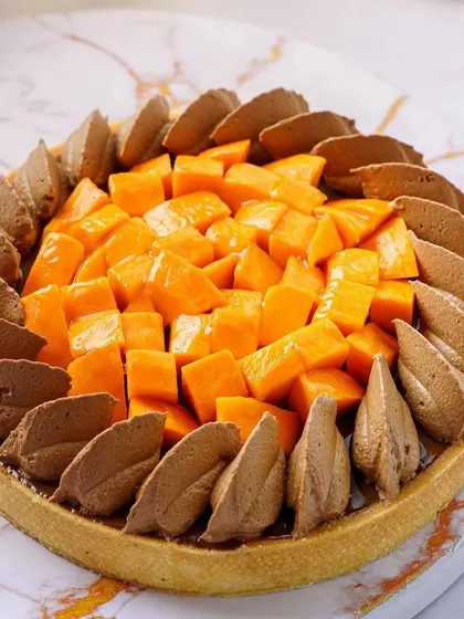 A stunning chocolate and mango tart. The contrast between the dark chocolate and bright mango makes for a visually striking photograph, shot on a marble background to add a touch of elegance.