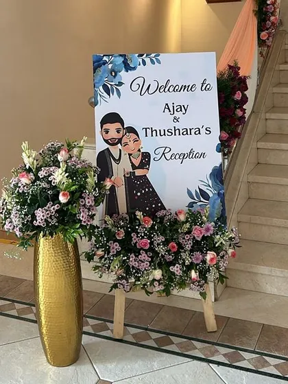 We design personalized welcome signs that capture the couple's personality, like this one featuring a cute caricature. The sign is placed on an easel and surrounded by flowers that match the reception's decor.