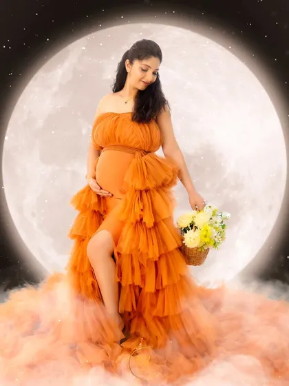 A whimsical, moon-themed maternity portrait. The mother-to-be sits on a cloud of orange tulle in a matching gown, holding a basket of flowers in front of a giant moon.