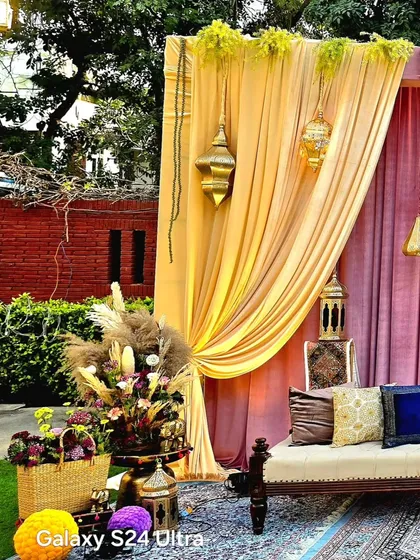 A cozy seating area at a Sangeet event, featuring a comfortable sofa with colorful cushions, set against a backdrop of pink and yellow drapes. The Moroccan lanterns and pampas grass arrangements add a touch of boho elegance.