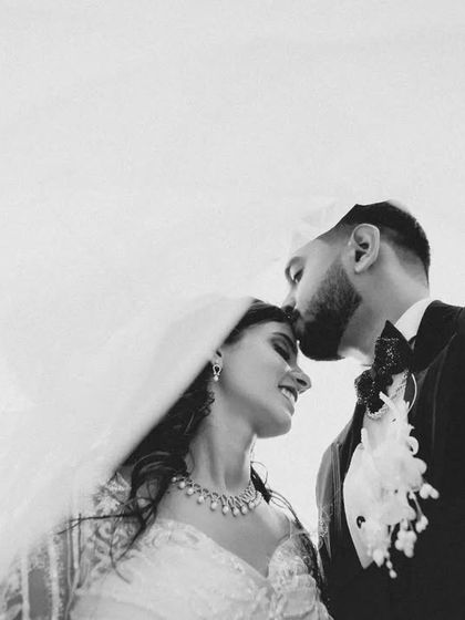 A tender black and white portrait of the groom kissing the bride's forehead under her veil. The high-key background keeps the focus entirely on their connection.