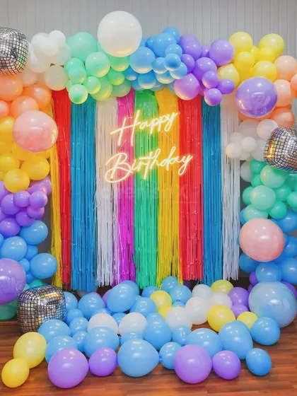 A burst of color for a fun and vibrant birthday party. This photo backdrop features a rainbow-colored fringe wall, a 'Happy Birthday' neon sign, and an explosion of multi-colored balloons, complete with disco balls for extra sparkle.