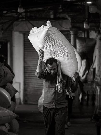 A laborer carries a heavy sack on his shoulder through the corridors of the Khari Baoli spice market. The soft, directional light models his form and conveys the physical effort of his work.