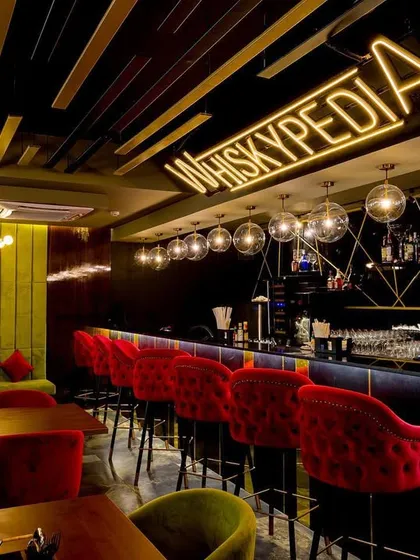 A full view of the 'Whiskypedia' bar, where every detail is curated to enhance the classic whiskey bar experience. The tufted red bar stools and olive green banquette seating add a touch of old-world luxury.