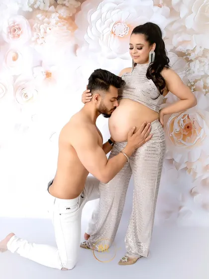 A modern and stylish couple's portrait. The partner kneels to kiss his partner's baby bump as she stands before a beautiful white floral wall.