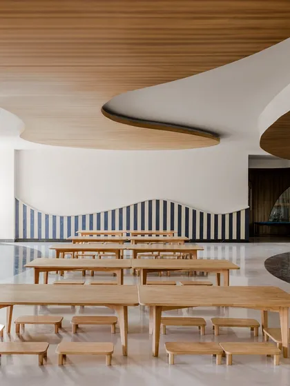 A dining or activity area at Vedanya School, featuring long wooden tables and benches. The curved ceiling and striped wall pattern continue the school's theme of fluid, imaginative design.