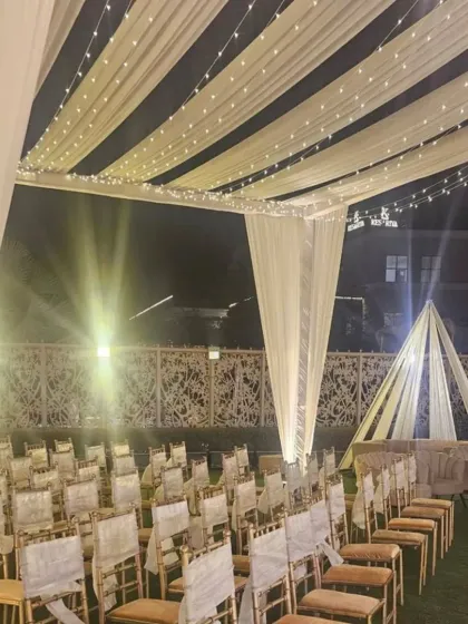 An elegant outdoor ceremony setup with a canopy of white drapes and fairy lights. The aisle is lined with chairs for guests, all facing a simple yet beautiful stage, creating a classic and romantic setting.