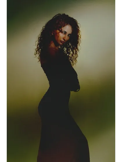A simple, elegant silhouette shot. The model is styled in a classic black dress, and the soft, greenish background light outlines her form and curly hair beautifully.