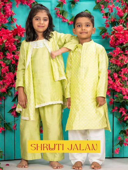 A brother and sister duo in a light lime green set. The boy's kurta has a subtle dot print, while the girl's outfit features a draped kurta with a delicate jacket, creating a fresh and modern festive look.