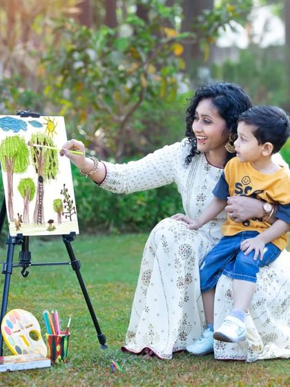 A mother and her young son are captured in a moment of shared creativity, painting together on an easel in a sun-dappled garden.