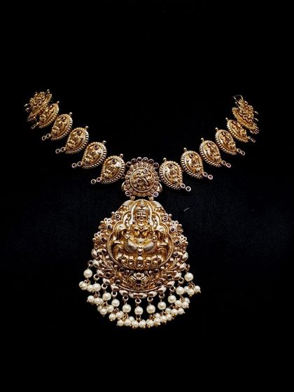 This is a classic mango-style temple necklace with a large, intricately carved Lakshmi pendant and pearl drops. The antique finish gives it a rich, heritage look.