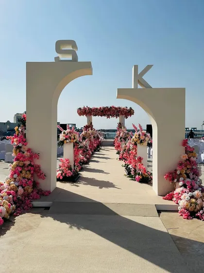 When the entrance archways on your wedding day look this grand and gorgeous. This modern arch, personalized with the couple's initials and adorned with pink flowers, sets the stage for a beautiful beachside ceremony. 🩷