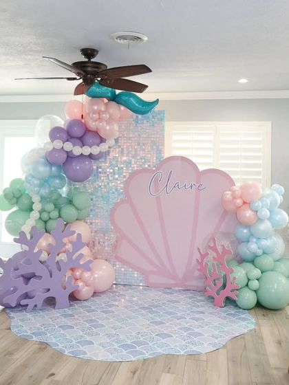 An "Under the Sea" mermaid party with a large pink seashell backdrop and a shimmering sequin panel. The scene is completed with coral props and a pastel balloon garland.